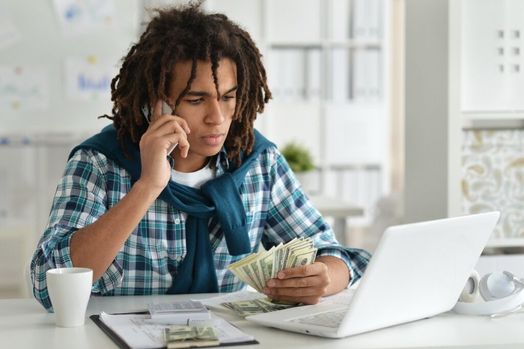 young afro man holding pile of money and using phone near laptop